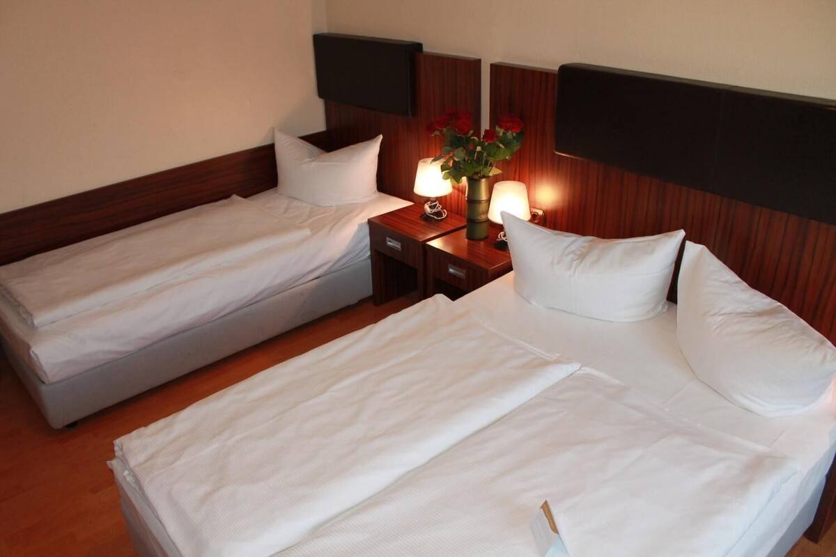 Une chambre d’hôtel avec deux lits simples, une table de chevet avec une lampe et un vase de fleurs, et des têtes de lit en bois.