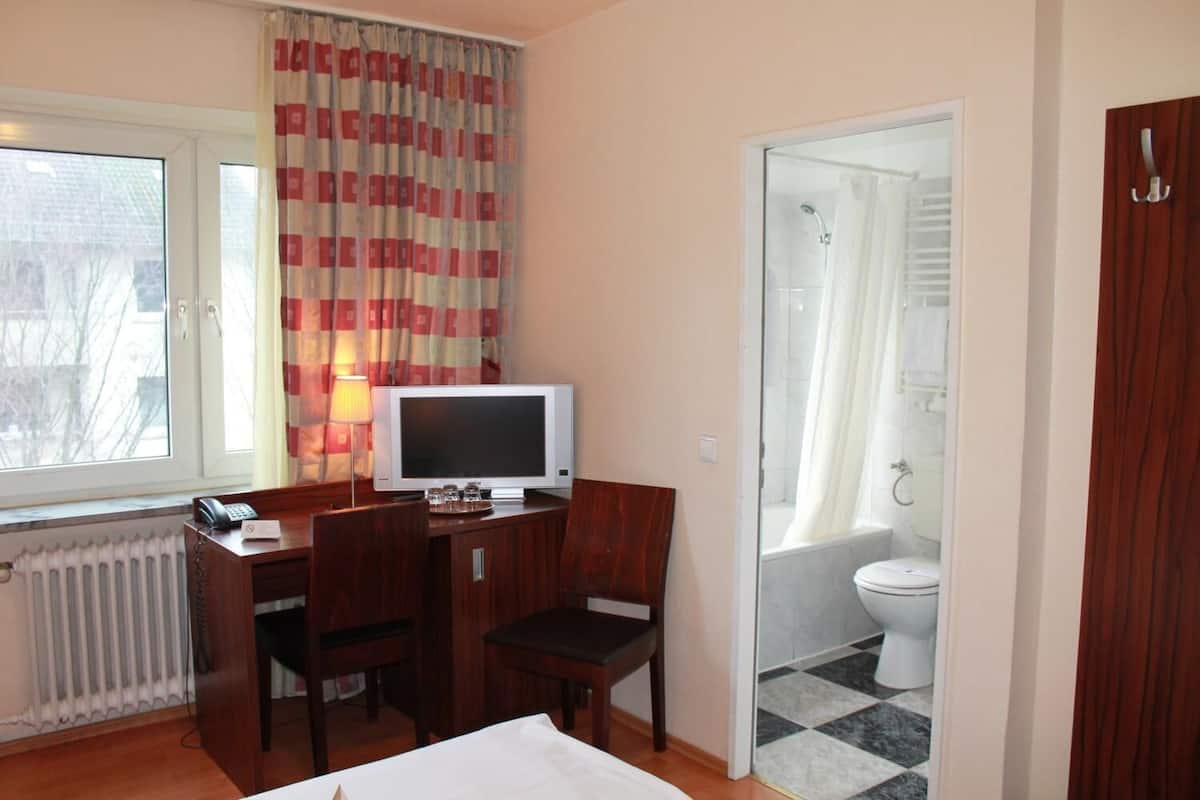Une chambre d’hôtel avec un bureau en bois, une télévision, une chaise, un radiateur, une fenêtre avec des rideaux et une salle de bain avec une douche et des toilettes.