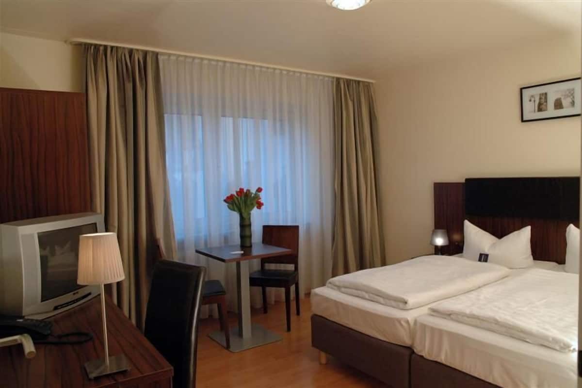 Une chambre d’hôtel avec un lit, un bureau avec une lampe, une chaise, un téléviseur et une fenêtre avec des rideaux.