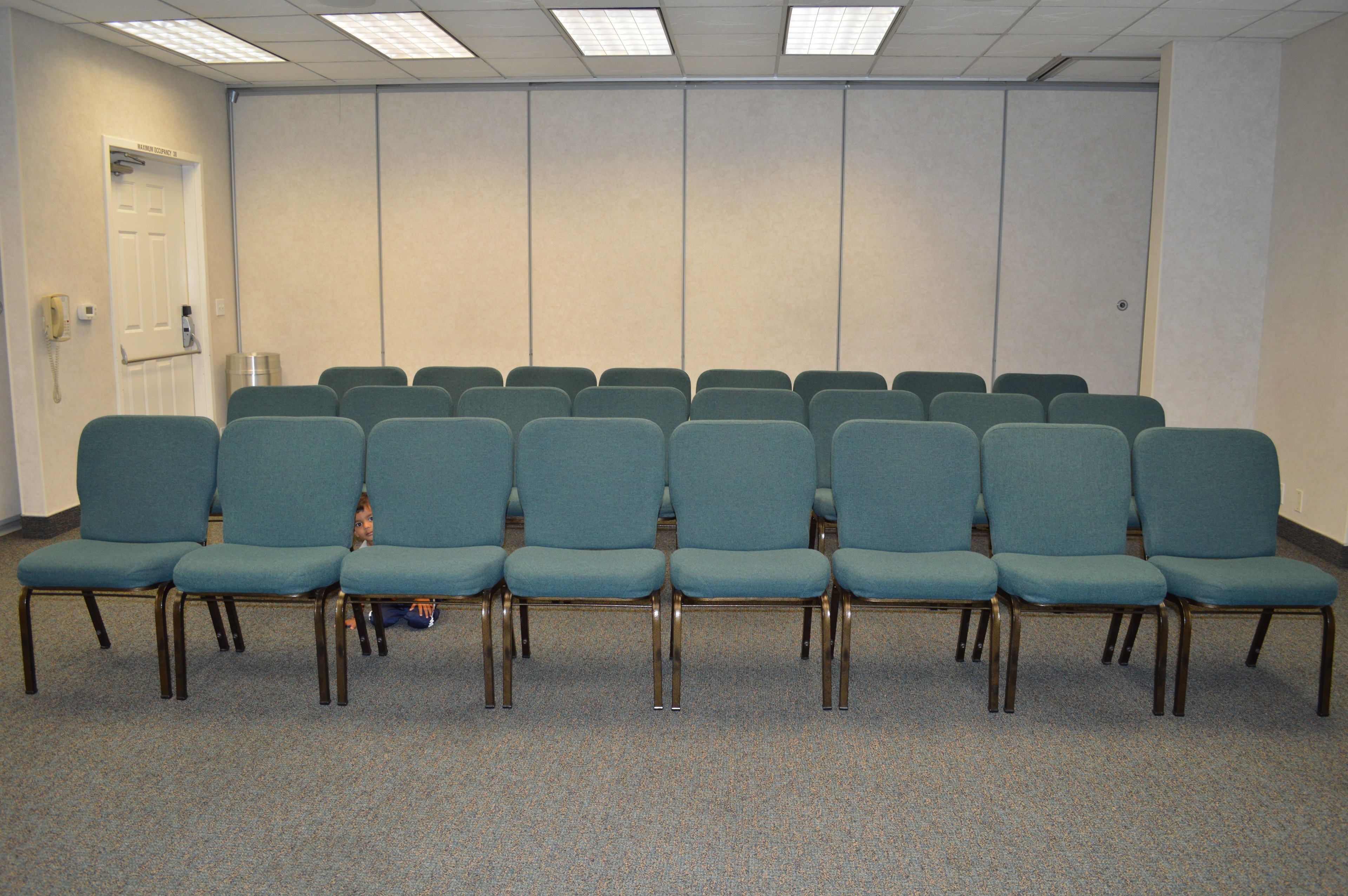 A conference room with rows of teal chairs, a white door, and a carpeted floor.