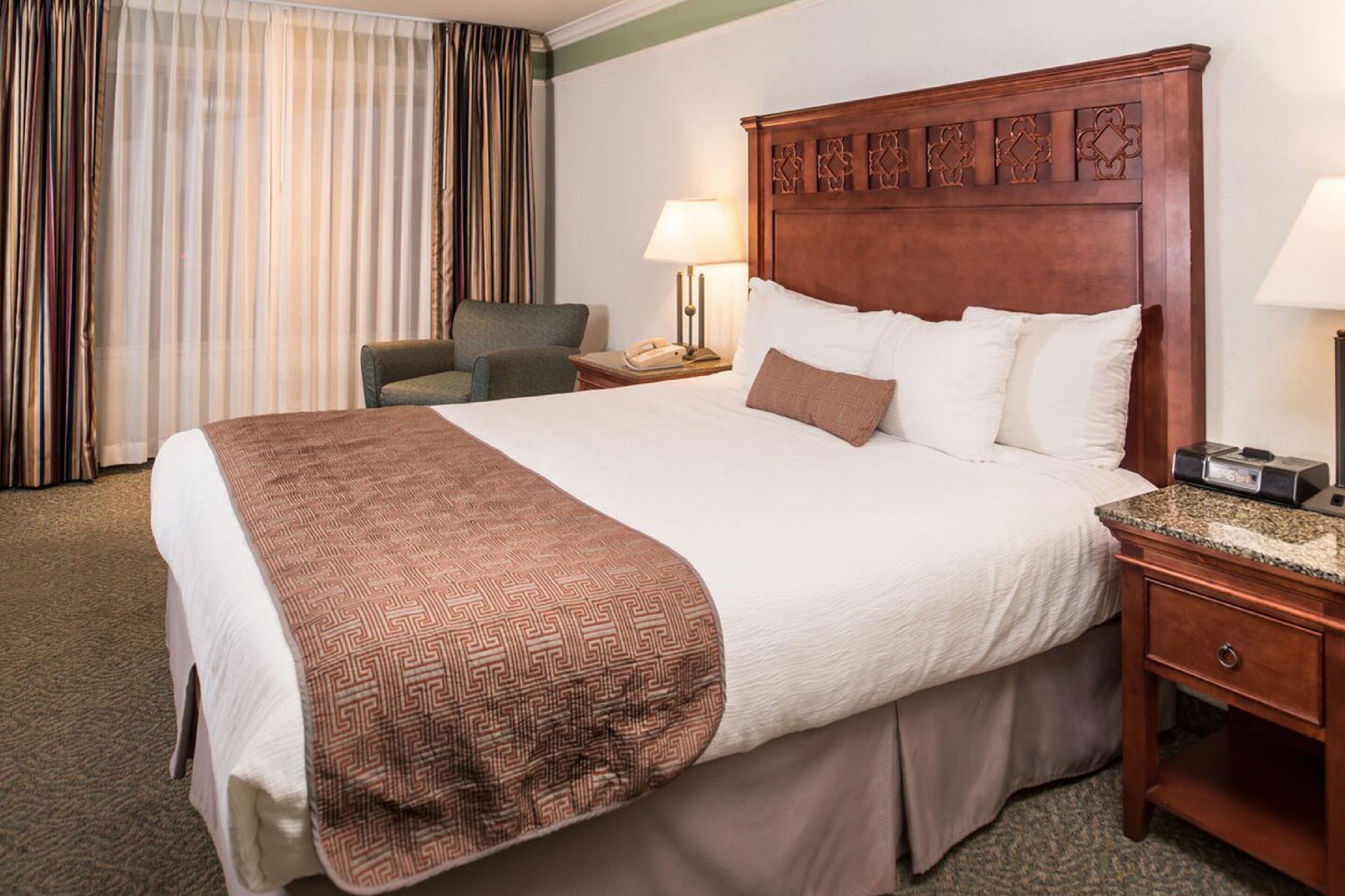 Una habitación de hotel con una cama grande, un cabecero de madera, un escritorio con lámpara, una silla y una ventana con cortinas.