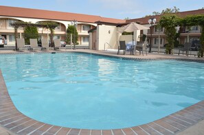 A swimming pool with lounge chairs and umbrellas, surrounded by a brick border and a building with a tiled roof.