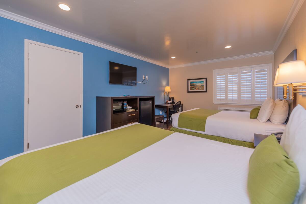 A hotel room with two beds, a desk, a TV, and a window with blinds.