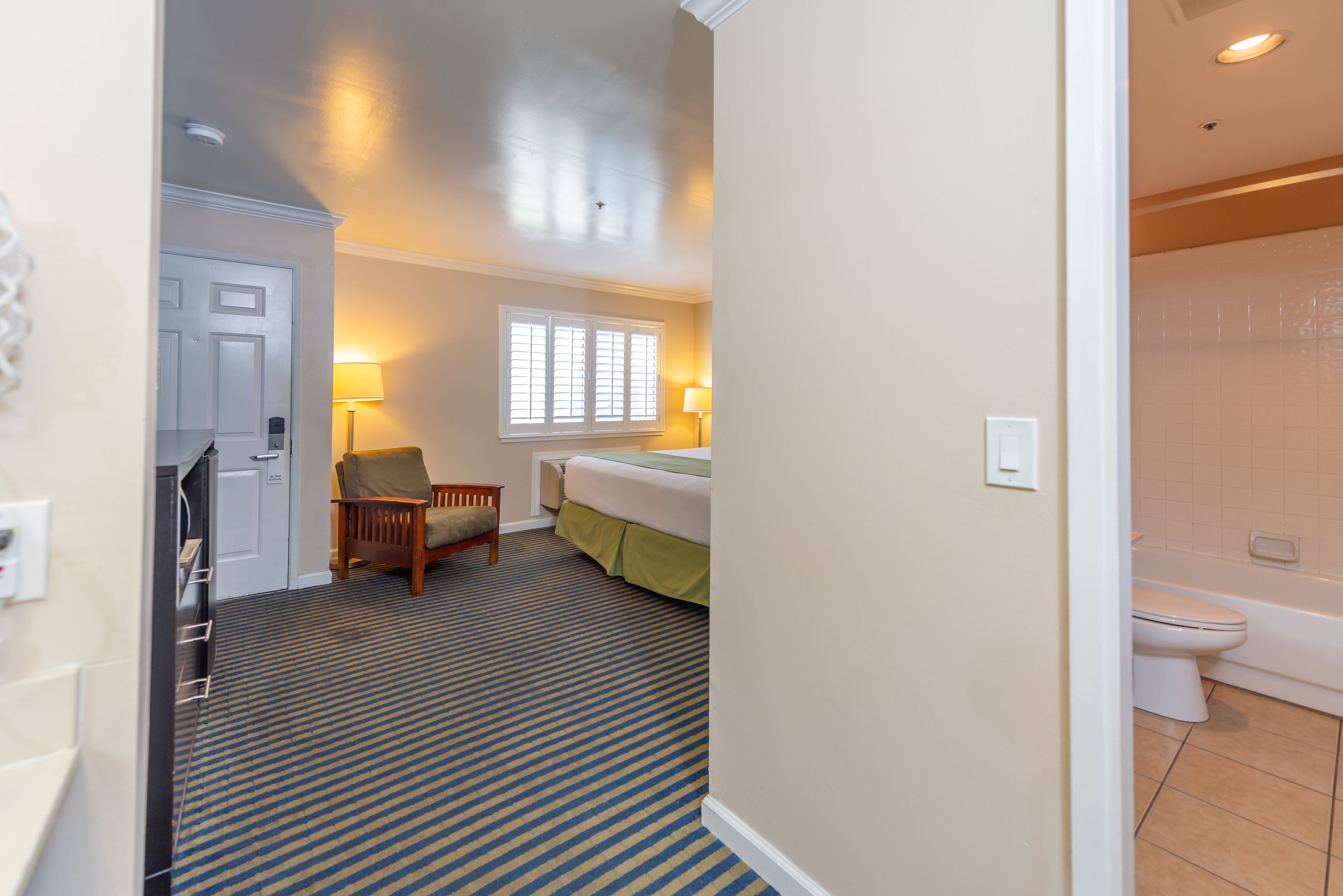 A hotel room with a bed, a chair, a window with blinds, a lamp, and a bathroom with a toilet and bathtub.