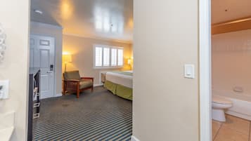 A hotel room with a bed, a chair, a window with blinds, a lamp, and a bathroom with a toilet and bathtub.
