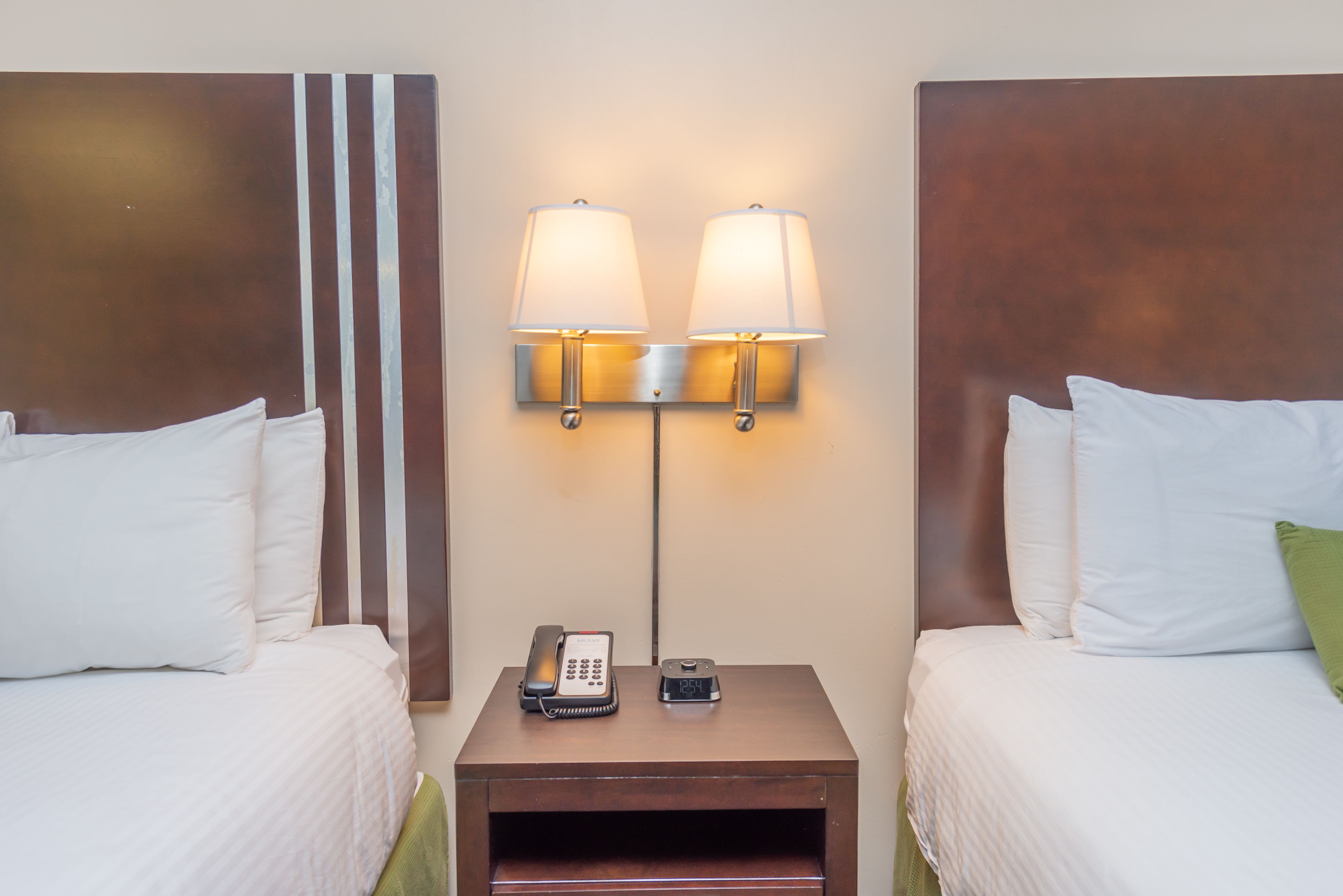 Una habitació d'hotel amb dos llits, una tauleta de nit amb un telèfon i un rellotge digital, i una làmpada penjada a la paret.