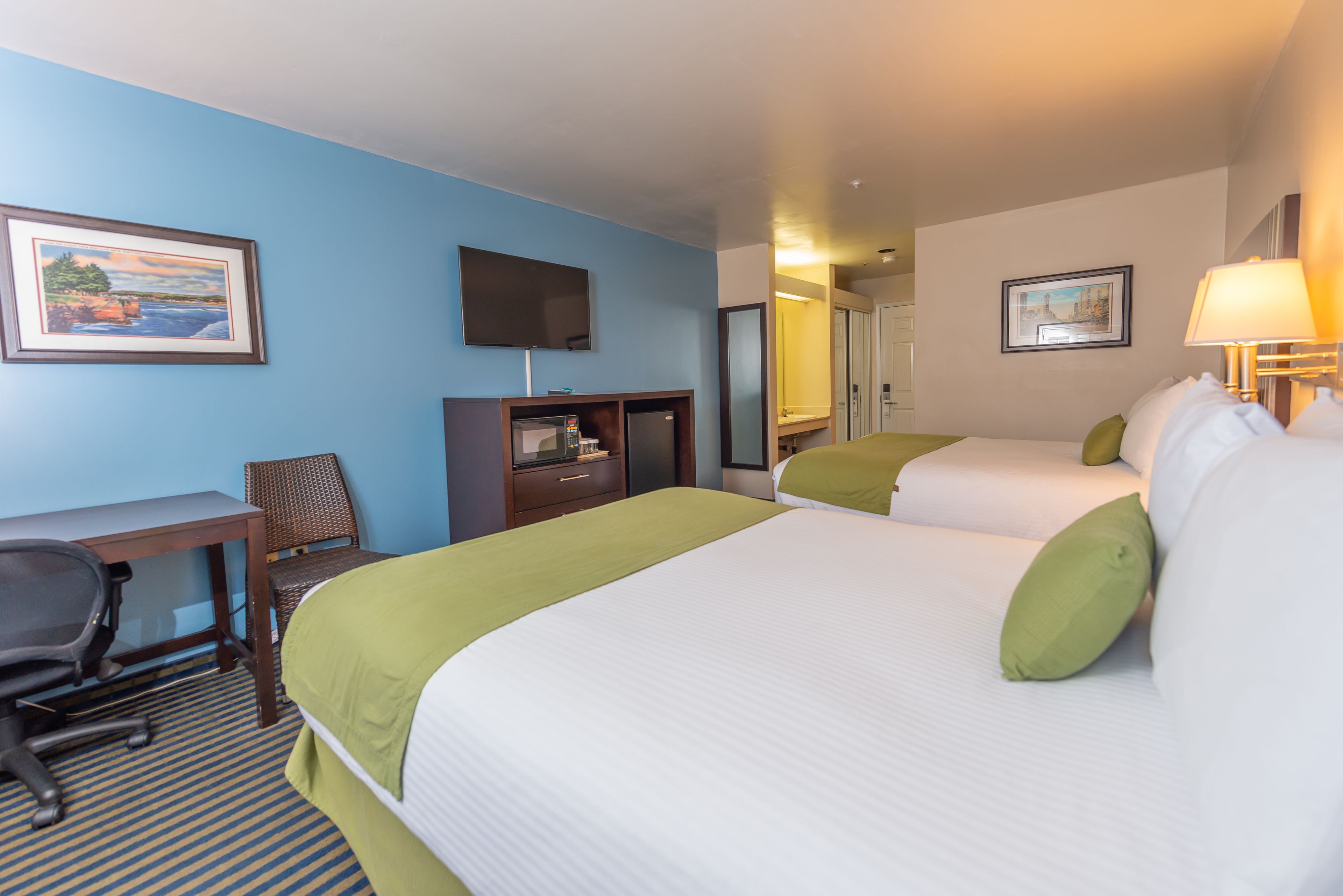 A hotel room with two beds, a desk, a chair, a TV, and artwork on the walls.