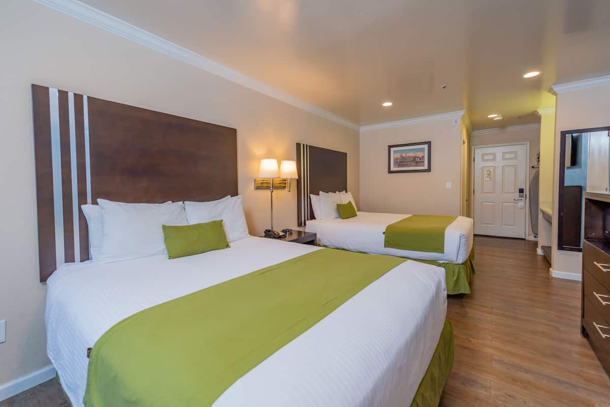 A hotel room with two beds, each with white bedding and green accents, a bedside table with lamps, and a door leading to a hallway.