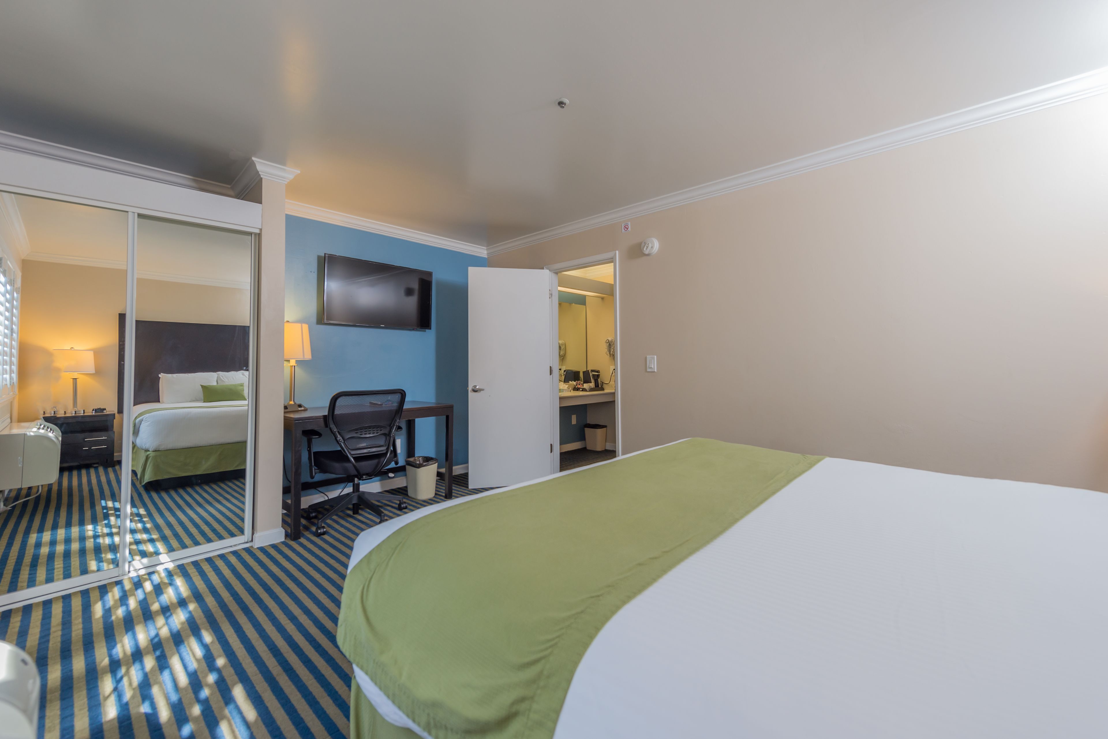 A hotel room with a bed, a desk, a chair, a TV, and a bathroom visible through an open door.