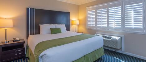 A hotel room with a large bed, two bedside tables, a nightstand, a window with blinds, and a wall-mounted air conditioner.
