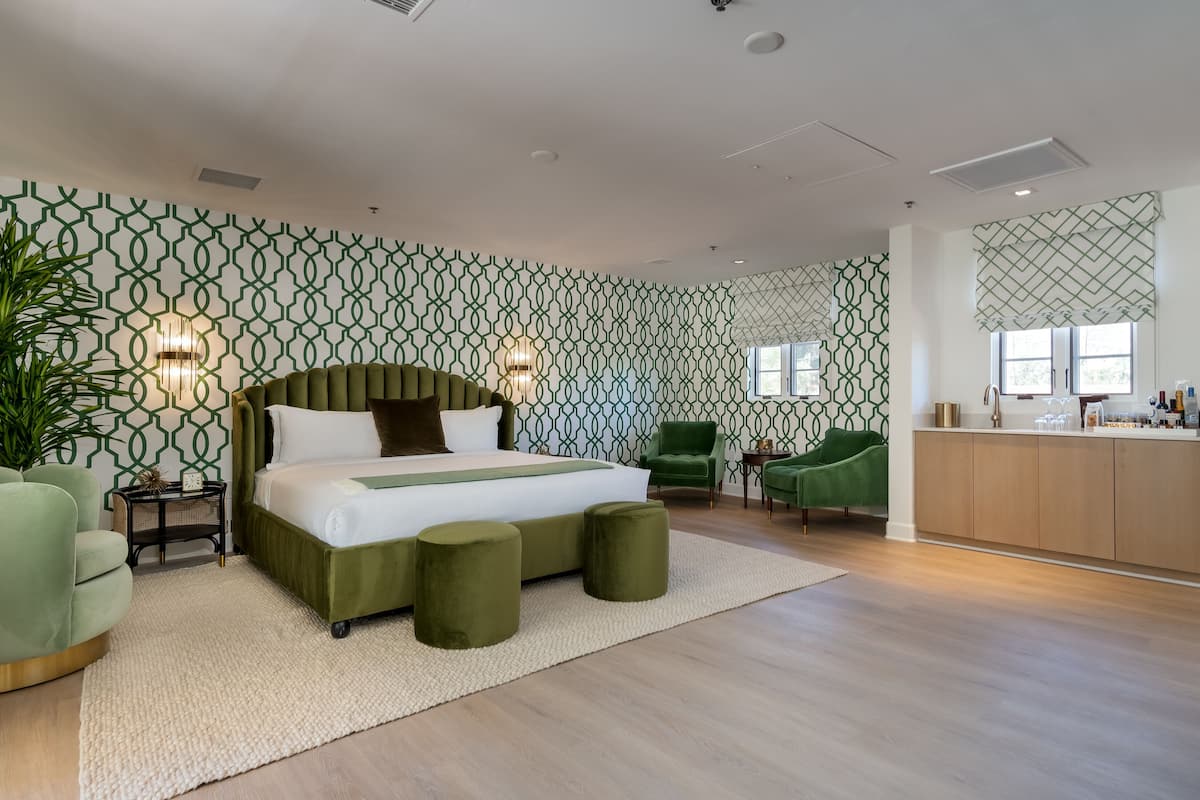 Un dormitorio moderno con una cama grande, sillas tapizadas de verde, una mesita auxiliar pequeña y una zona de cocina con un armario de madera.