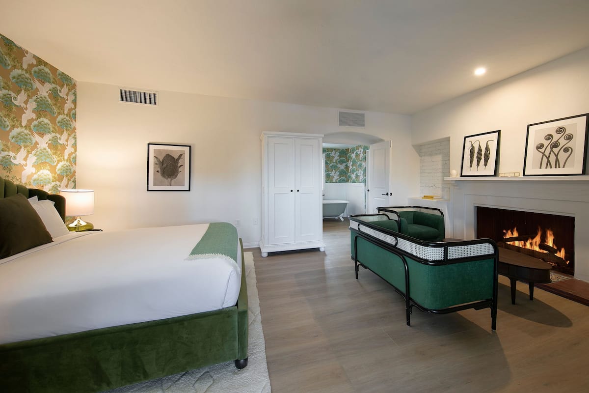 Un dormitorio con una cama, un sillón verde, una chimenea y una puerta que da a otra habitación.