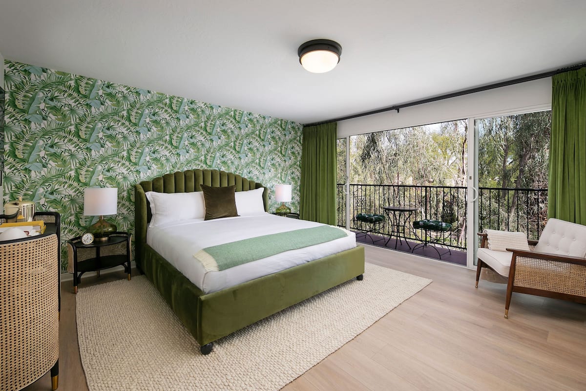Un dormitorio con una cama grande, un cabecero verde, una mesita de noche con lámpara, una silla de ratán y un balcón con vistas a árboles.