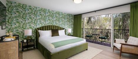 Une chambre avec un grand lit, une tête de lit verte, une table de chevet avec une lampe, un fauteuil en rotin et un balcon avec vue sur les arbres.