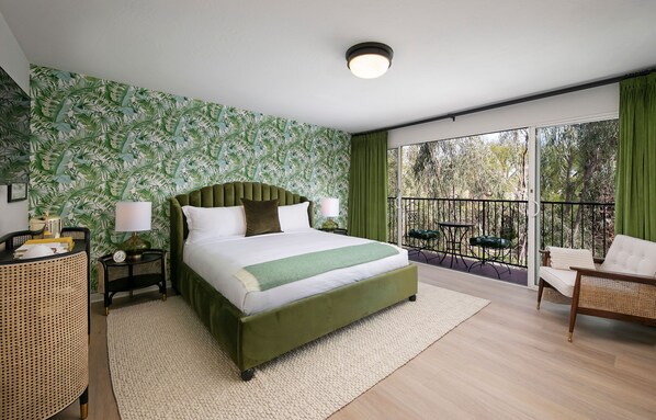A bedroom with a large bed, a green headboard, a bedside table with a lamp, a rattan chair, and a balcony with a view of trees.