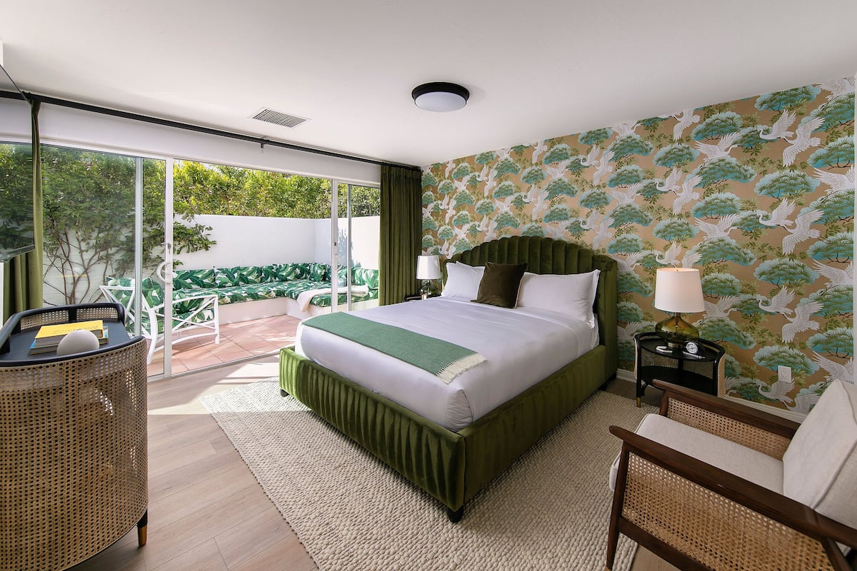 Habitación de hotel con una cama grande, cabecera verde, una mesita de noche con lámpara y vistas al exterior a través de una puerta corredera.