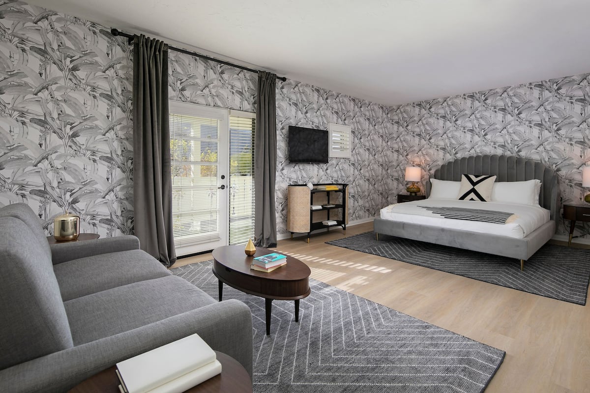 Un dormitorio moderno con un sofá gris, una cama con cabecero gris, una mesita redonda pequeña y una estantería.