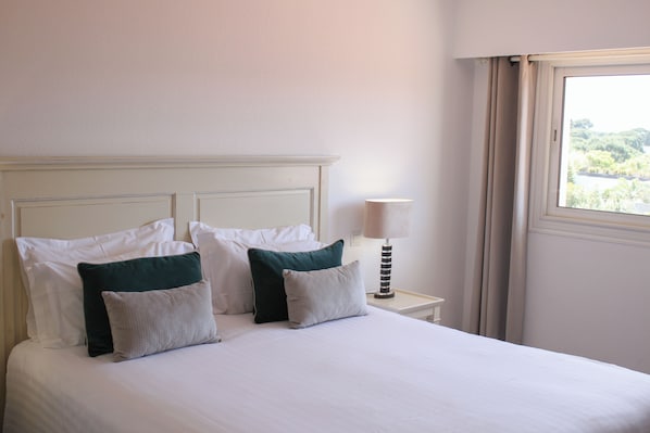 A neatly made bed with white and green pillows, a bedside lamp, and a window with a view of greenery.