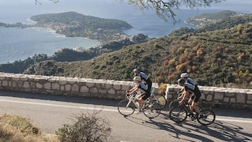 A coastal road with cyclists, stone walls, and a view of the sea and hills.