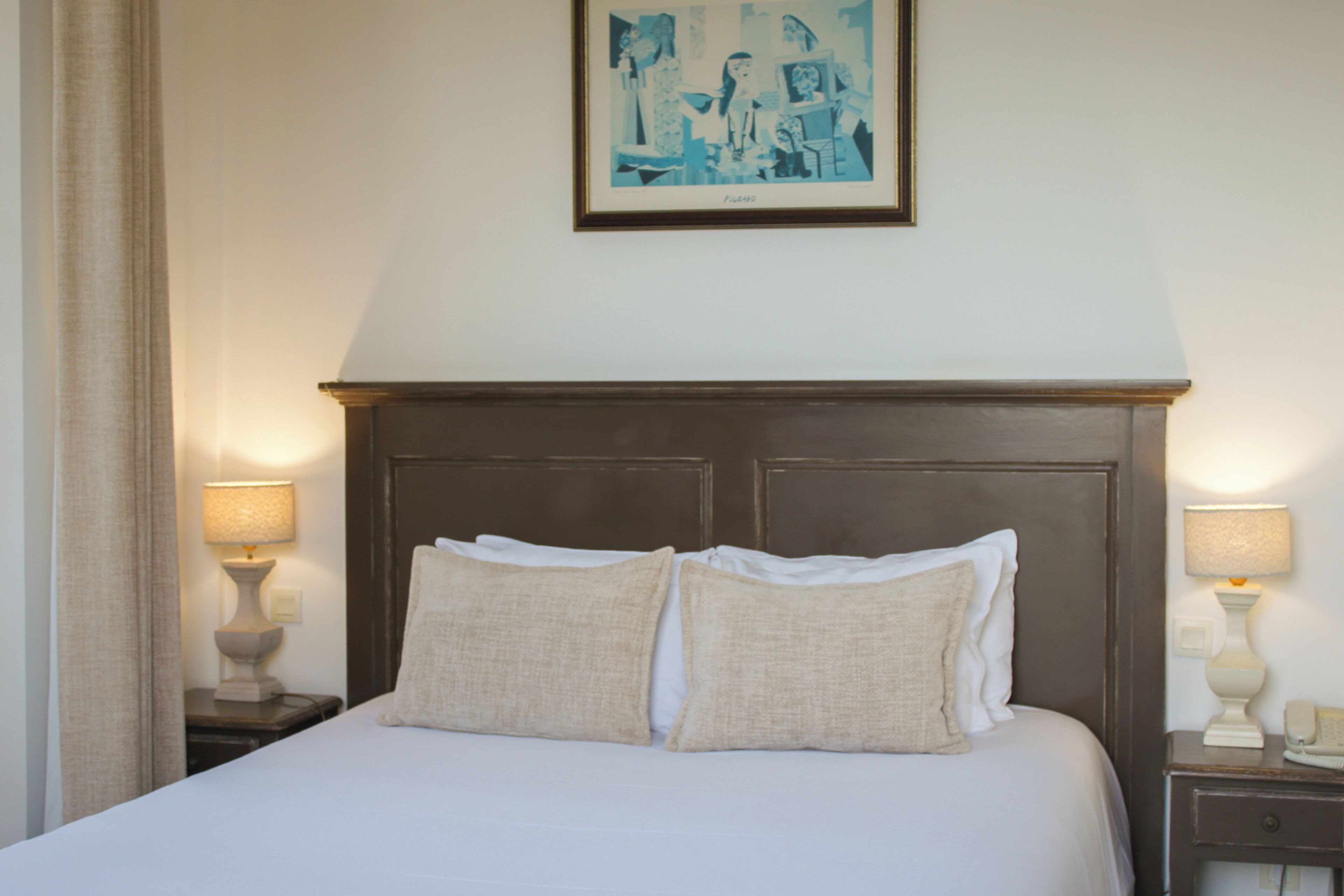 Een hotelkamer met een bed, twee nachtkastjes met lampen, een ingelijst schilderij aan de muur, en een gordijn aan de linkerkant.