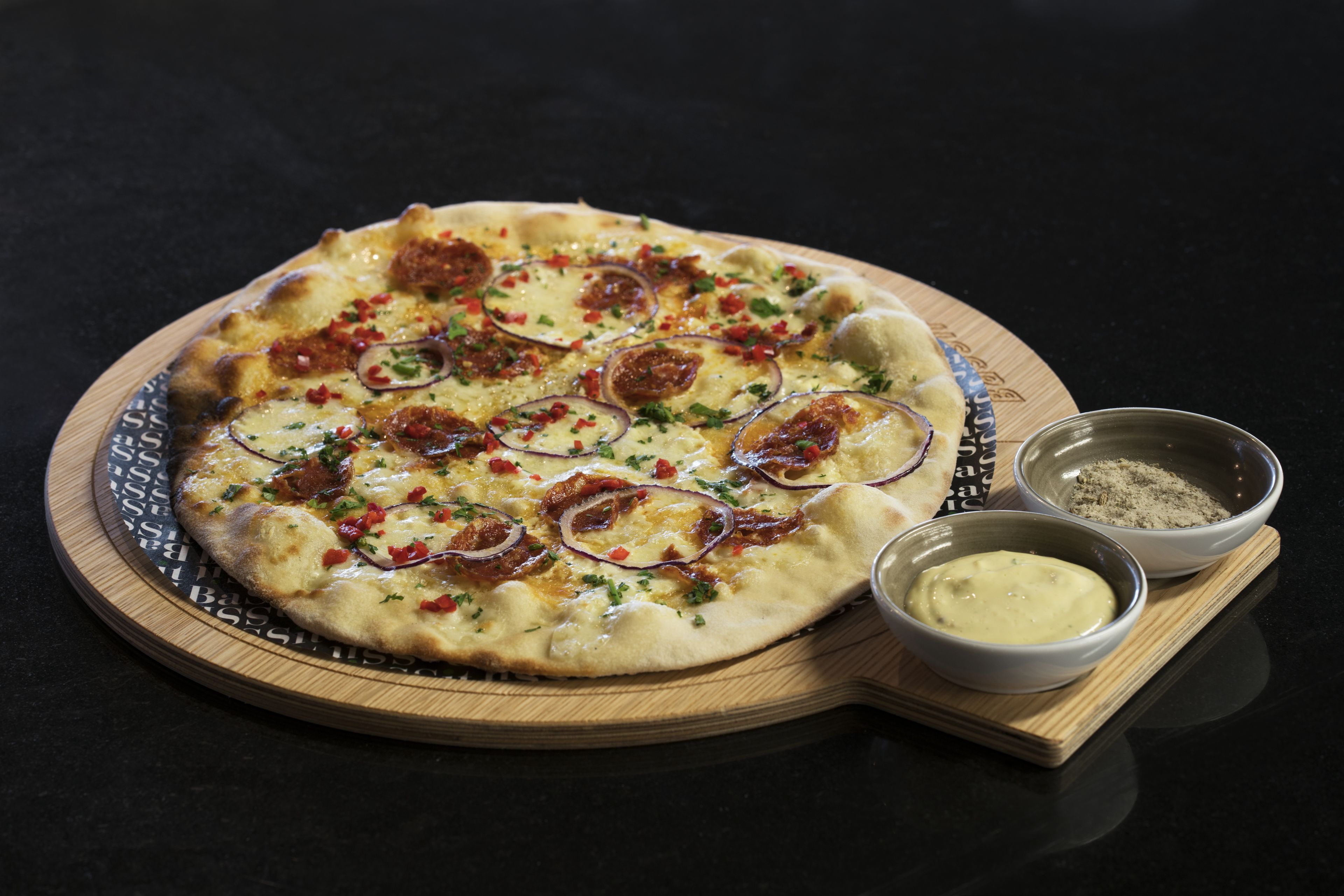 A pizza on a wooden board with two small bowls of dipping sauces.