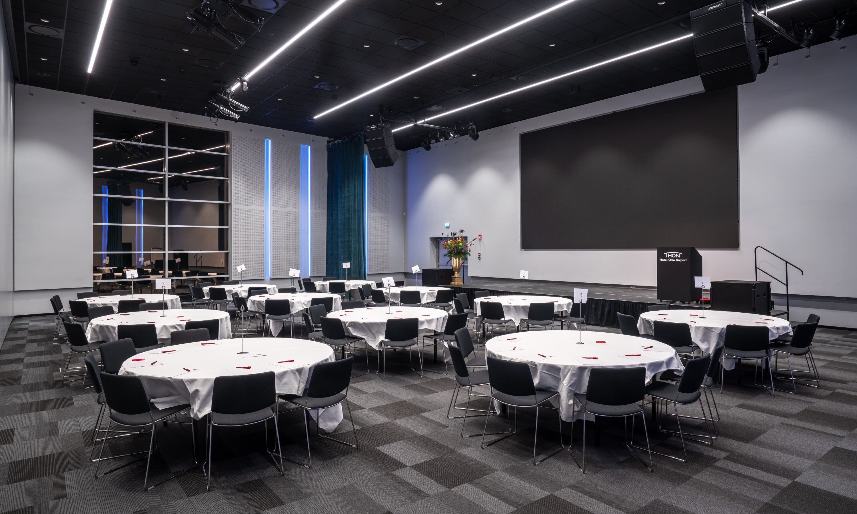 A conference room with multiple round tables, black chairs, and a large screen.