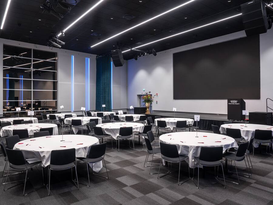 A conference room with multiple round tables, black chairs, and a large screen.