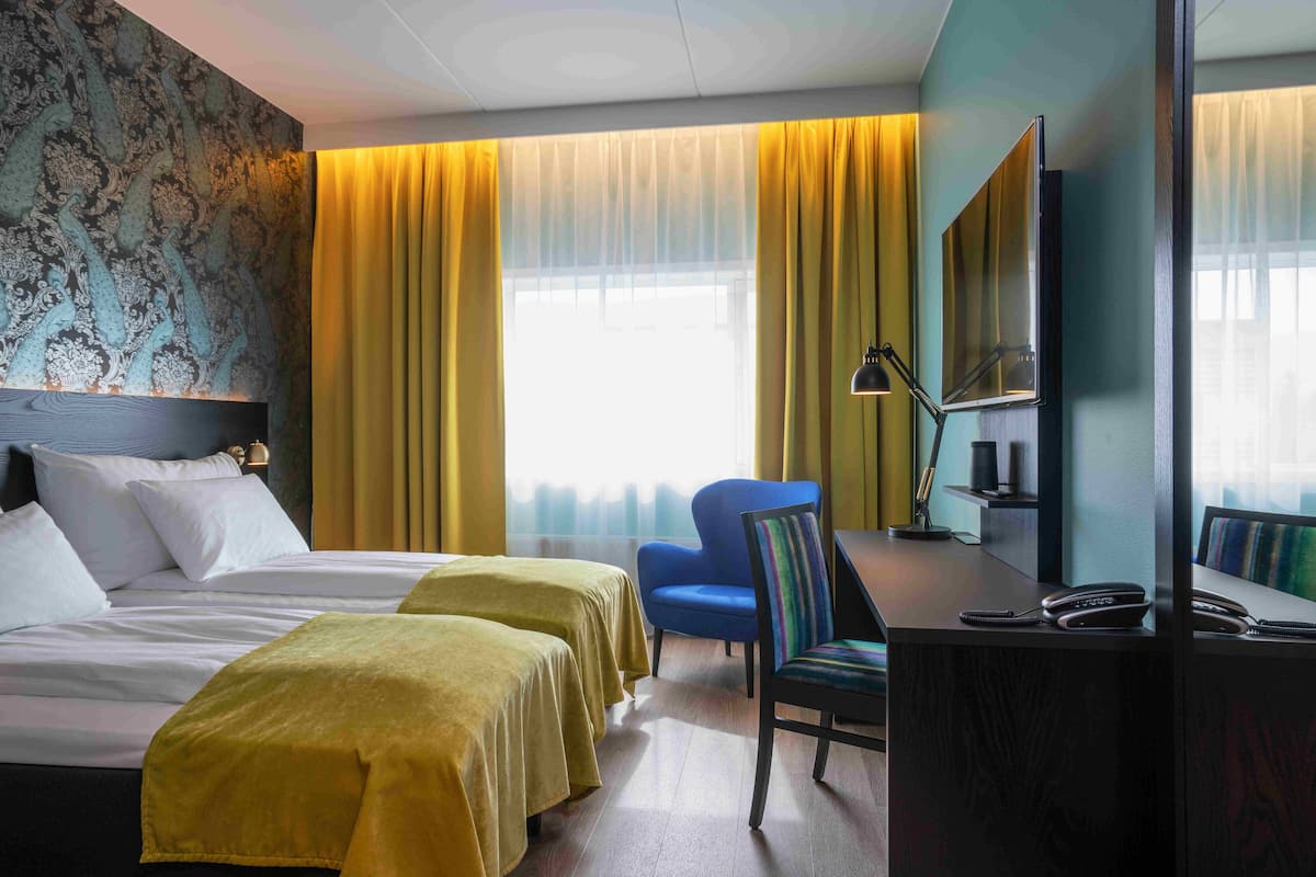 A hotel room with two beds, a desk, a chair, a mirror, and a window with yellow curtains.