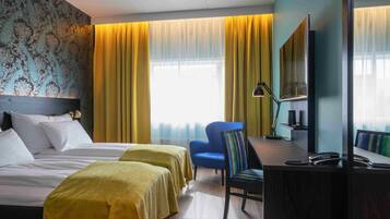 A hotel room with two beds, a desk, a chair, a mirror, and a window with yellow curtains.