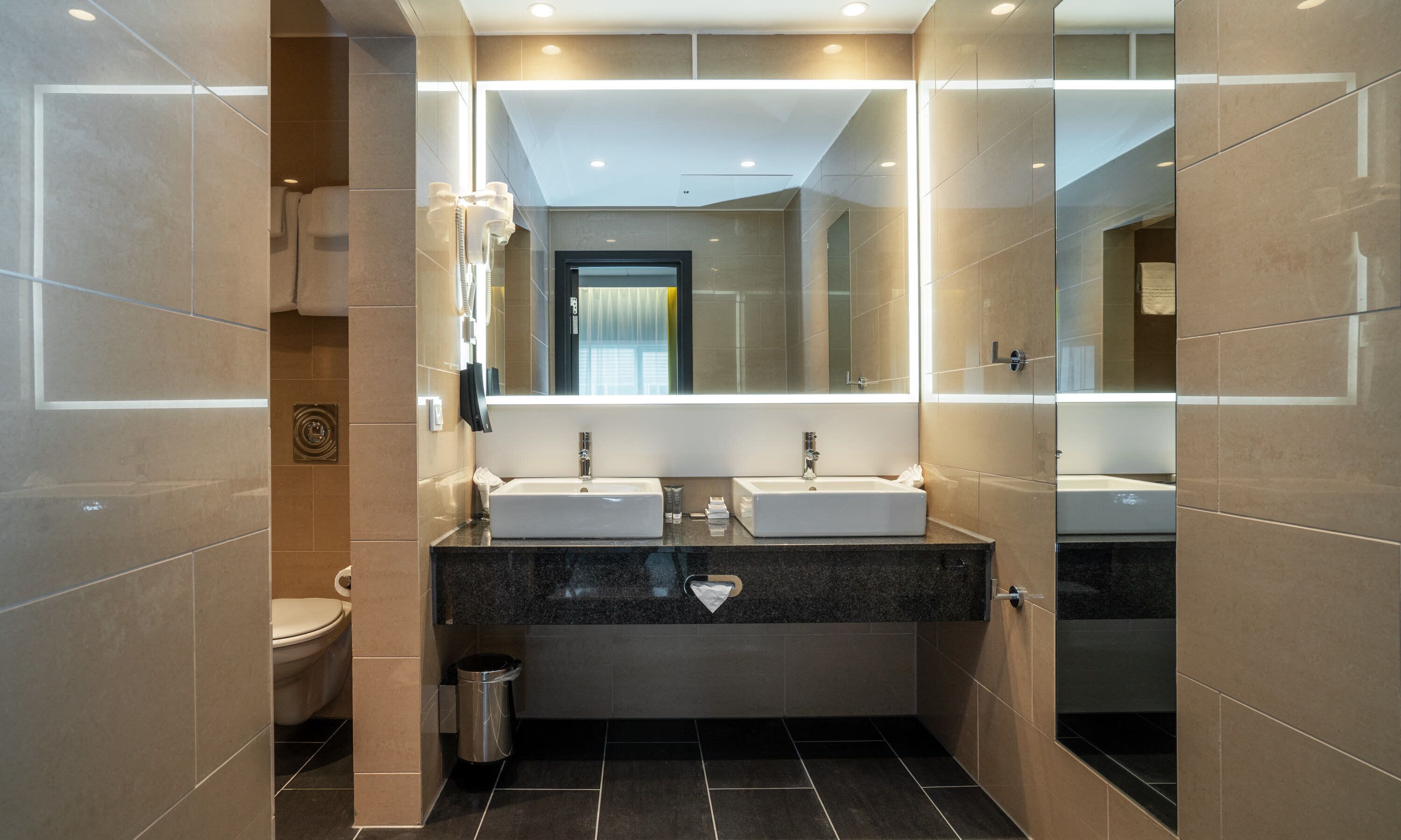 Un baño moderno con dos lavamanos, un inodoro y un cubo de basura.