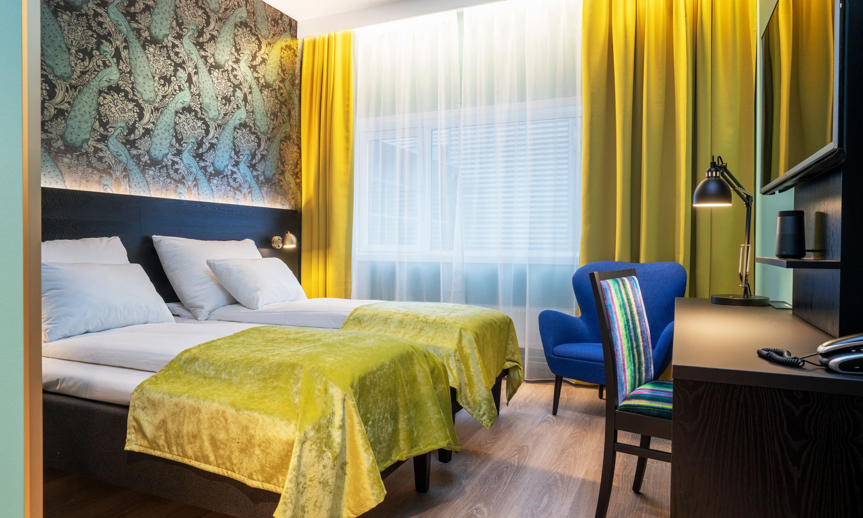 Habitación de hotel con dos camas, un escritorio con silla, una lámpara y un ventanal con cortinas amarillas.