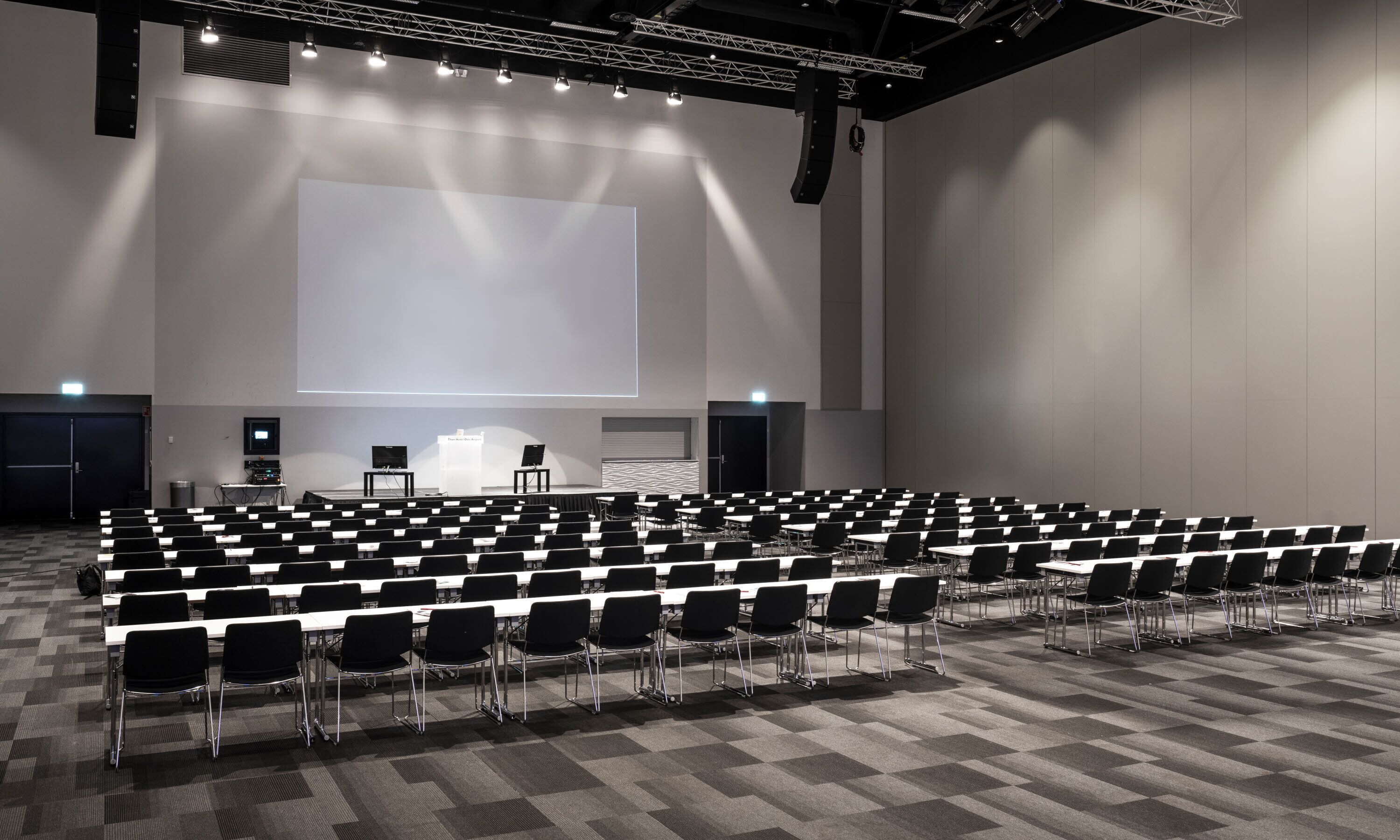 Ein großer Konferenzraum mit Reihen von Stühlen und Tischen, einer Bühne mit einem Bildschirm und einem Rednerpult.