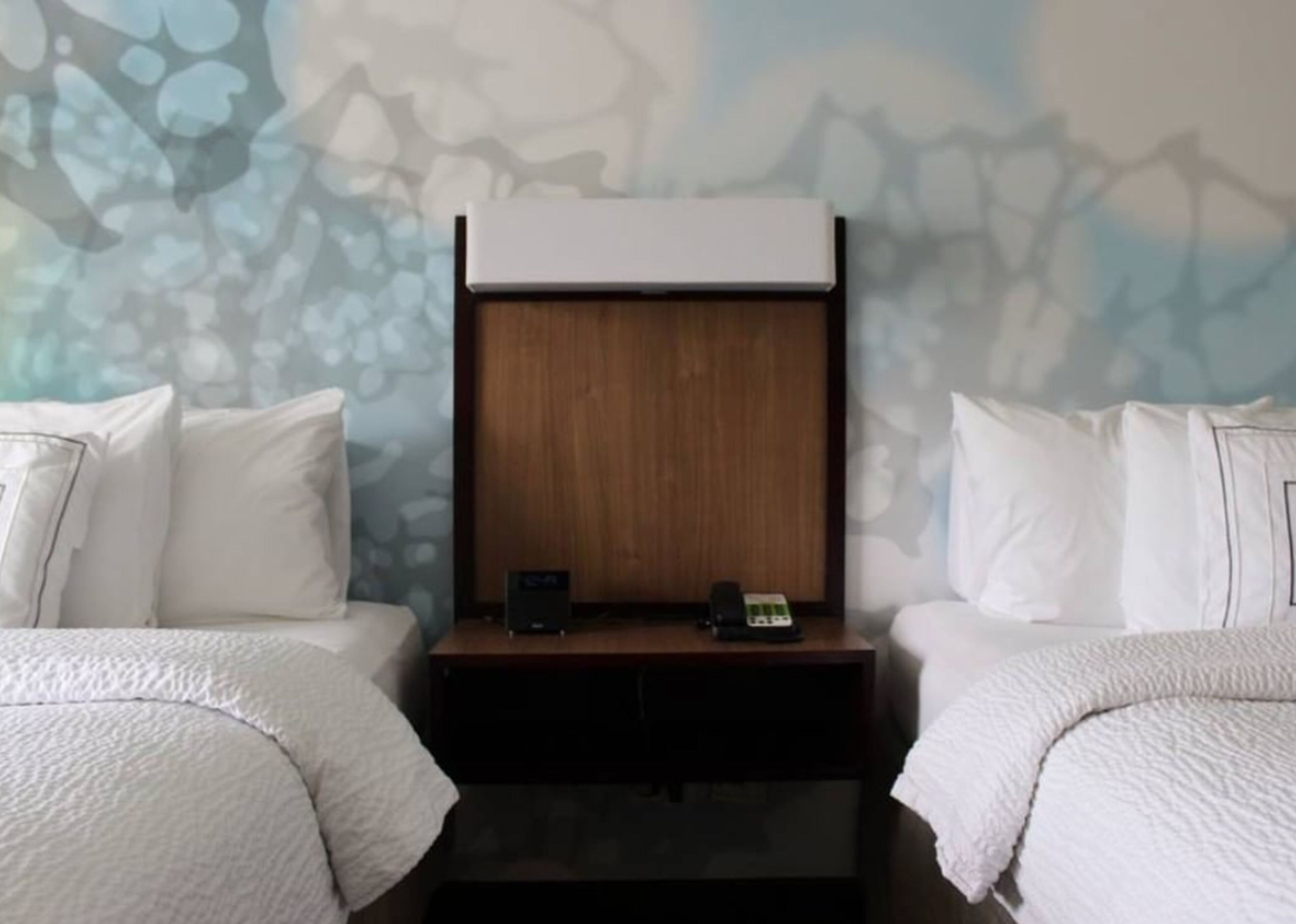 Quarto de hotel com duas camas, uma cabeceira de madeira, mesa de cabeceira com telefone e parede estampada com motivos florais.