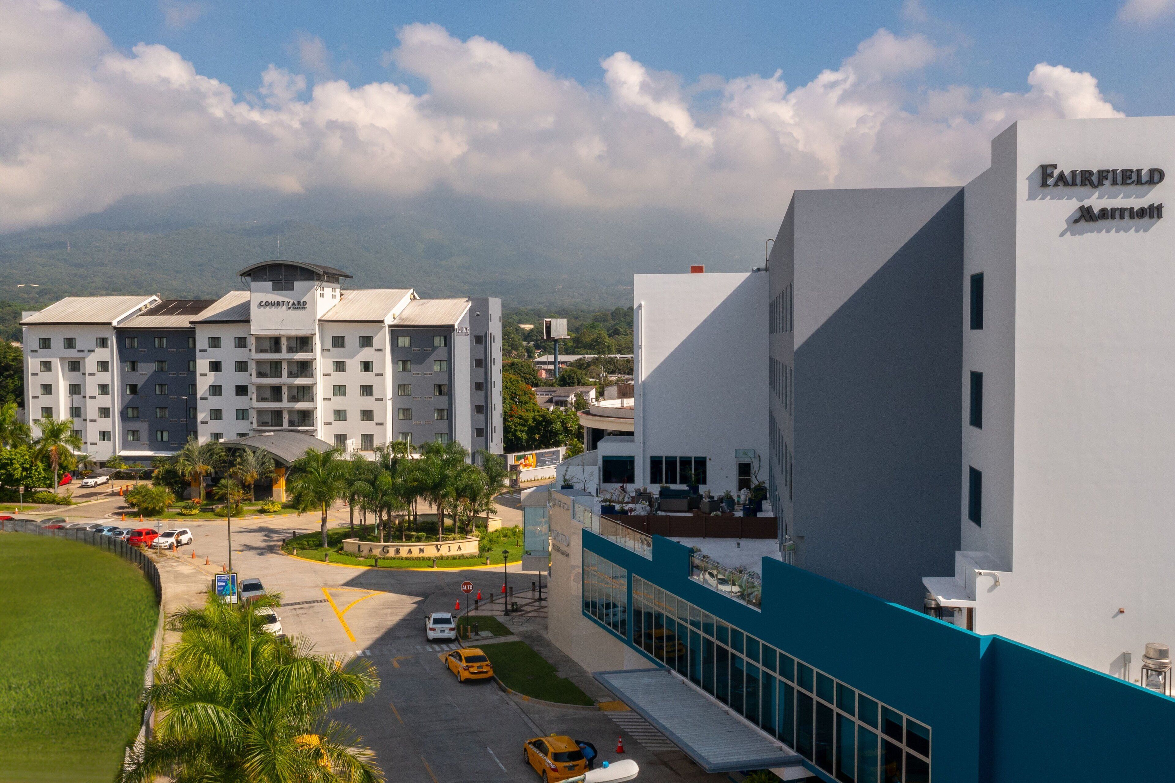 Un hotel Fairfield Marriott con diseño moderno, que presenta una fachada destacada en azul y blanco, un área de estacionamiento con varios autos, y vistas de edificios y zonas verdes circundantes.