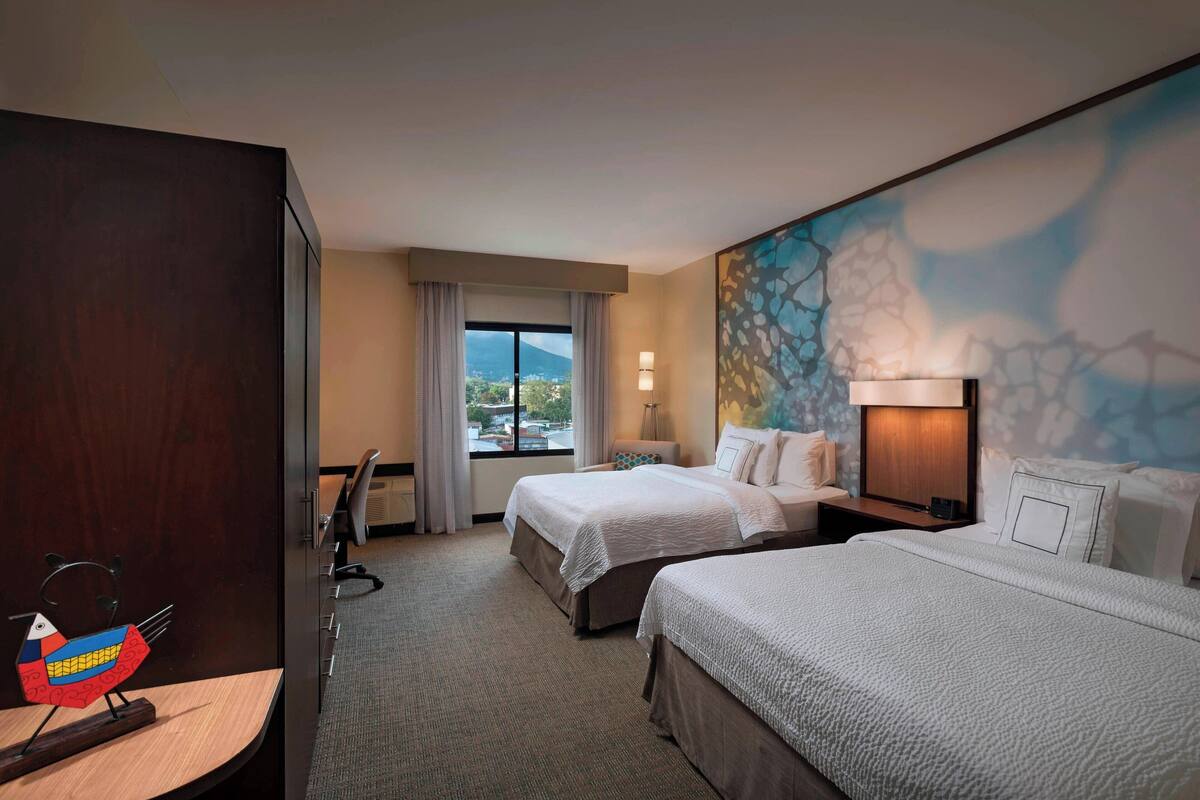 Ein Hotelzimmer mit zwei Betten, einem Schreibtisch, einem Stuhl, einer Lampe und Blick ins Freie.