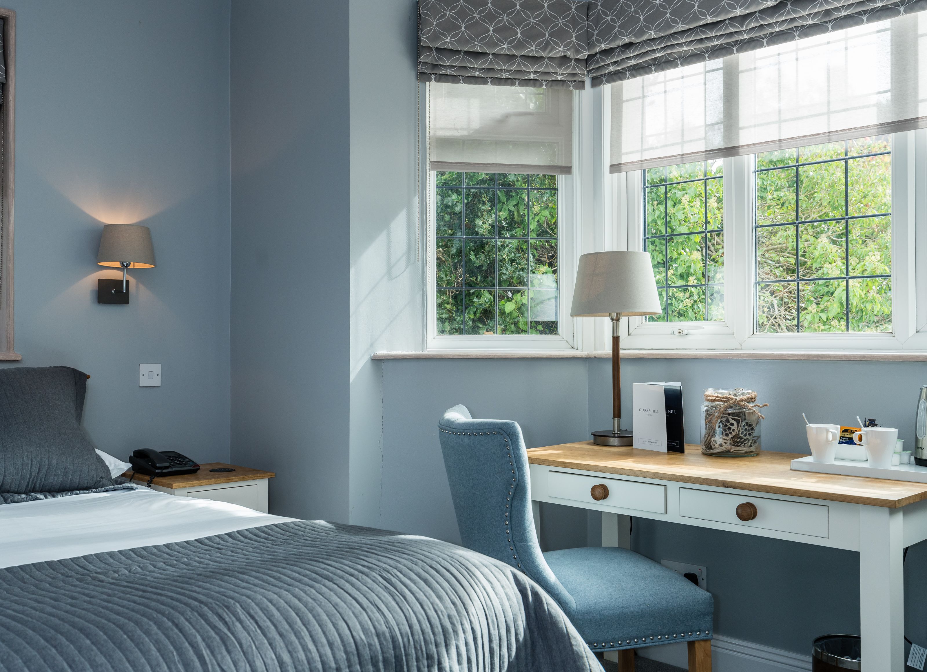 A bedroom with a bed, bedside lamp, desk, chair, and a window with a view of greenery.