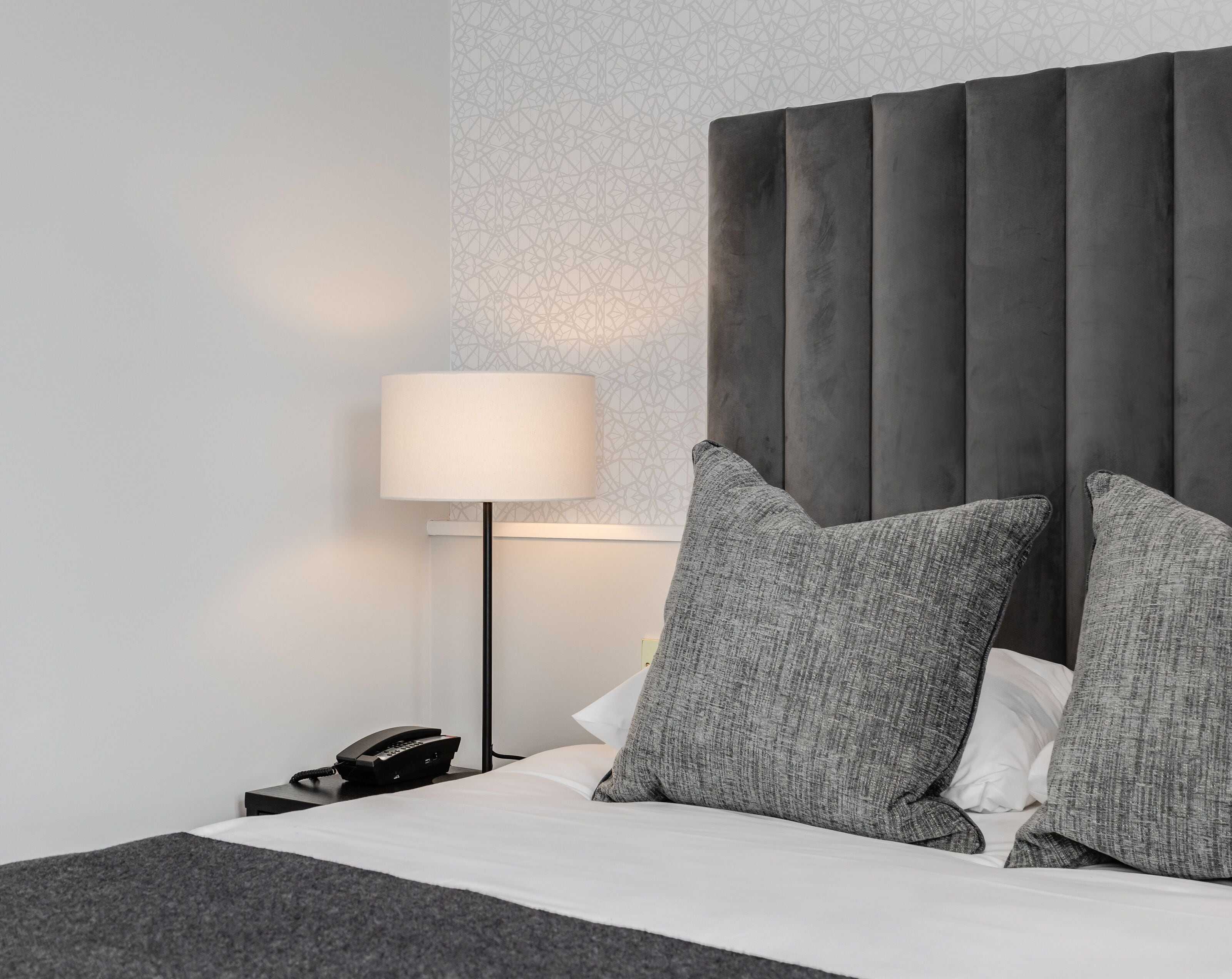 Ein modernes Hotelzimmer mit einem großen Bett, grauem Bettzeug, einem grauen Kopfteil, einer Nachttischlampe und einem Telefon auf dem Nachttisch.