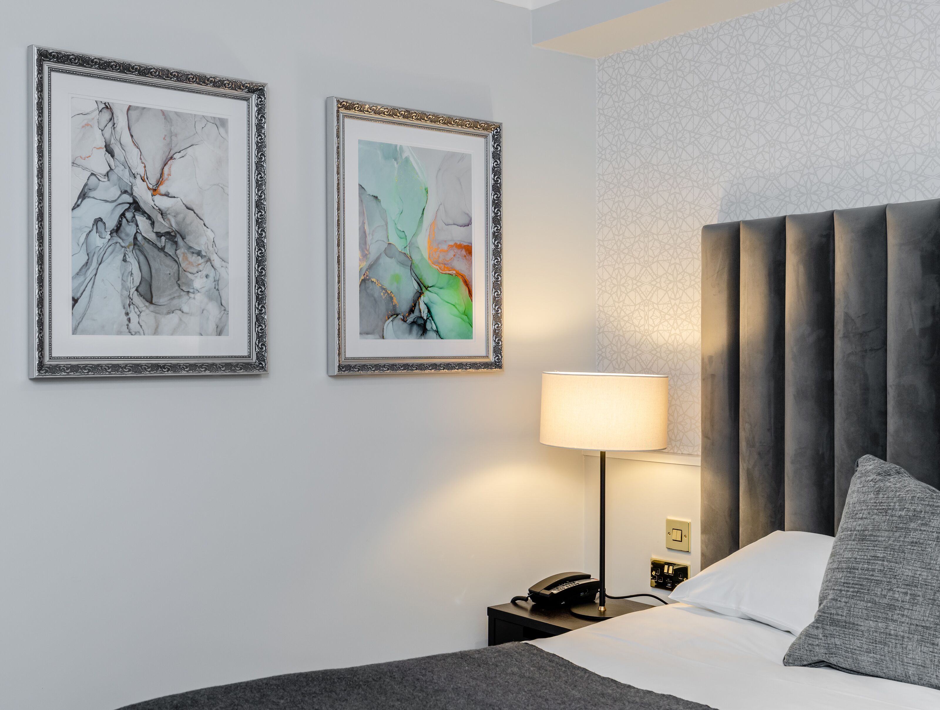 Quarto de hotel moderno com uma cama grande, uma mesinha de cabeceira com abajur e duas pinturas abstratas na parede.