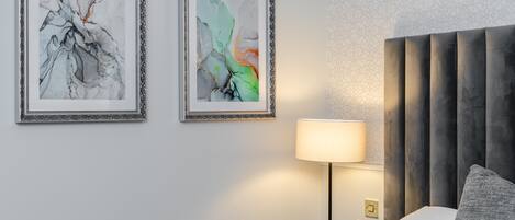 Quarto de hotel moderno com uma cama grande, uma mesinha de cabeceira com abajur e duas pinturas abstratas na parede.