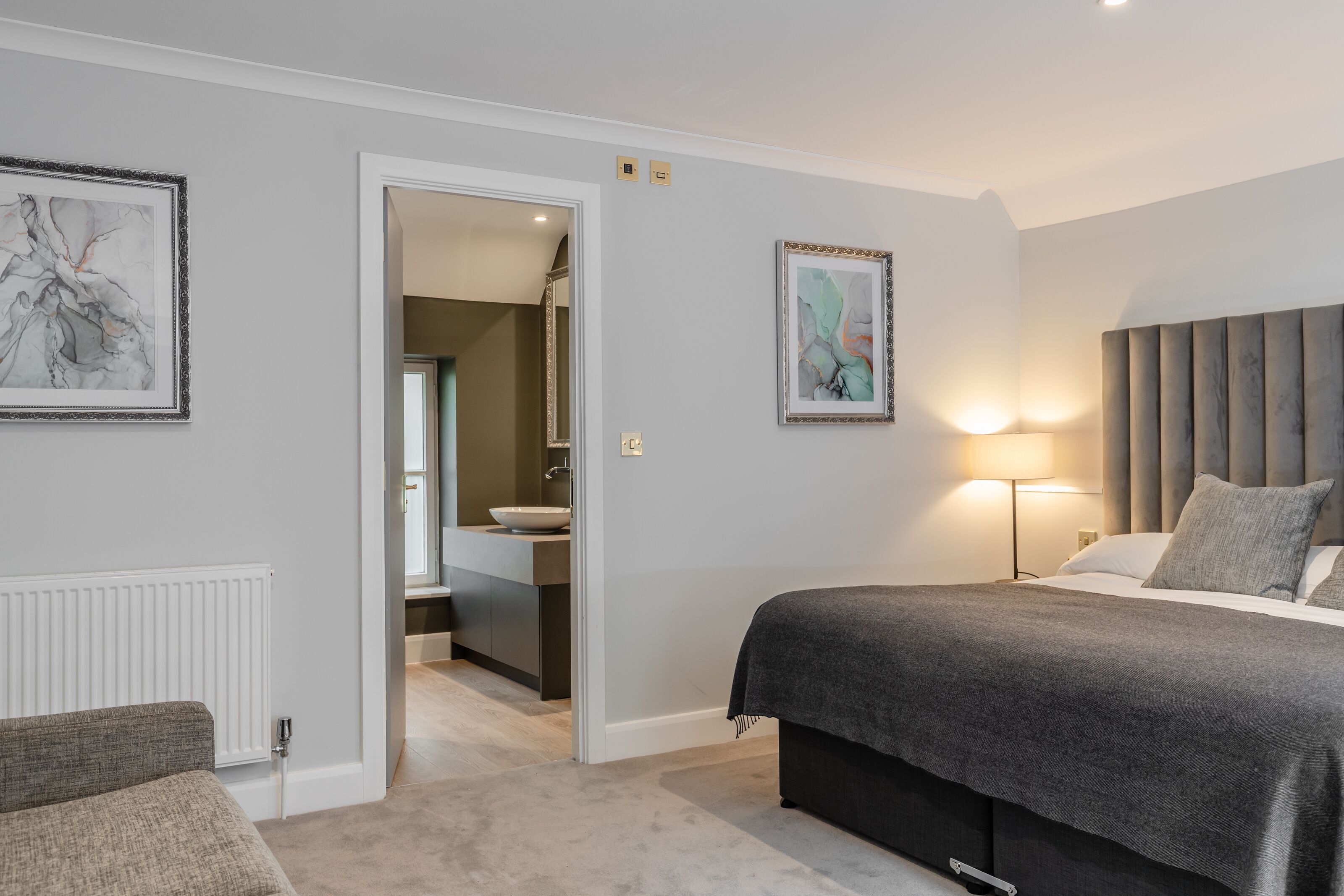 Un dormitorio moderno con una cama grande, una mesita de noche, una silla, un baño visible a través de una puerta abierta y dos obras de arte enmarcadas en las paredes.