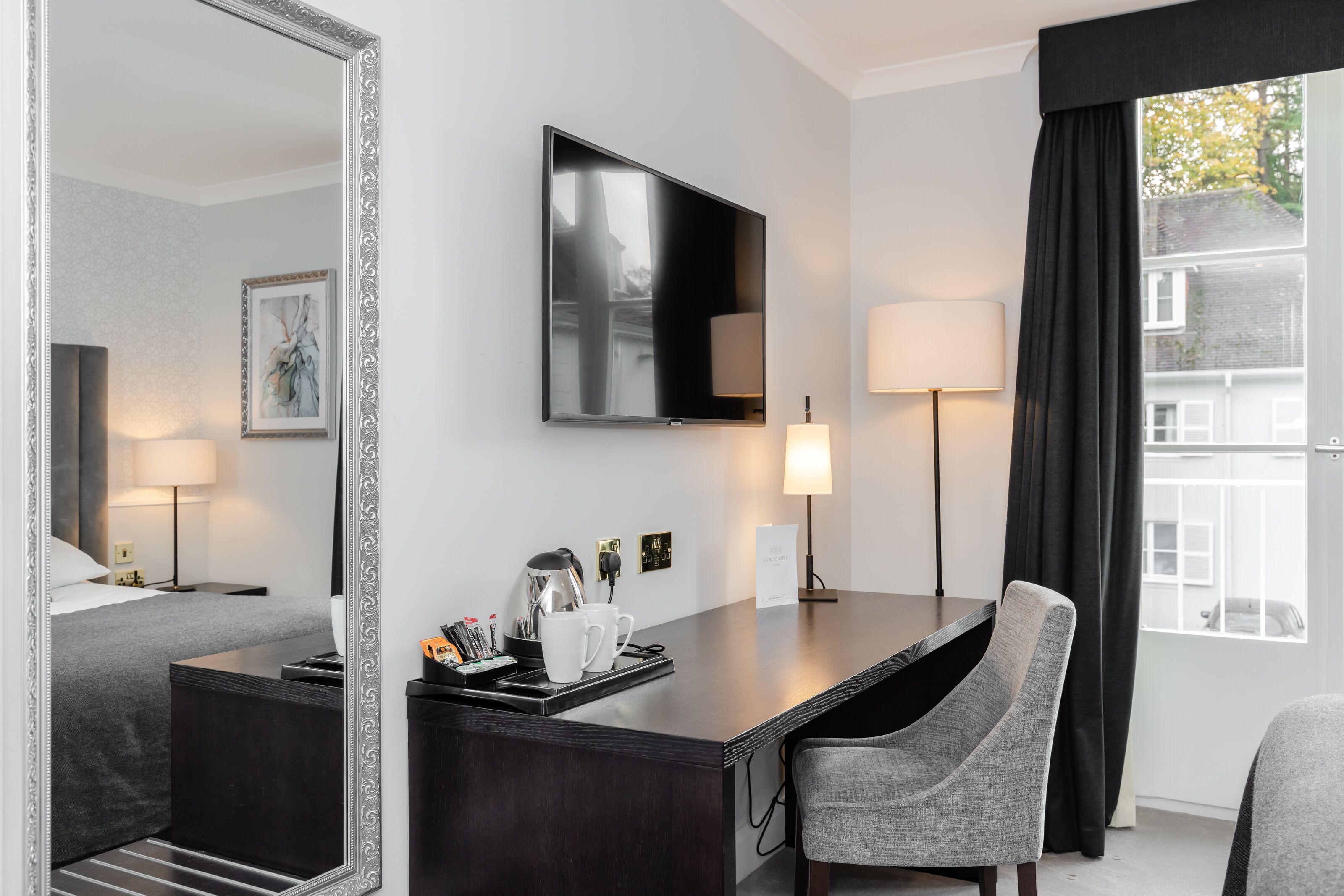 A hotel room with a bed, a desk with a chair, a flat-screen TV, a lamp, and a window with curtains.