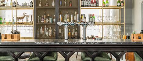 A bar area with a marble countertop, multiple shelves with bottles and glasses, and green upholstered chairs.