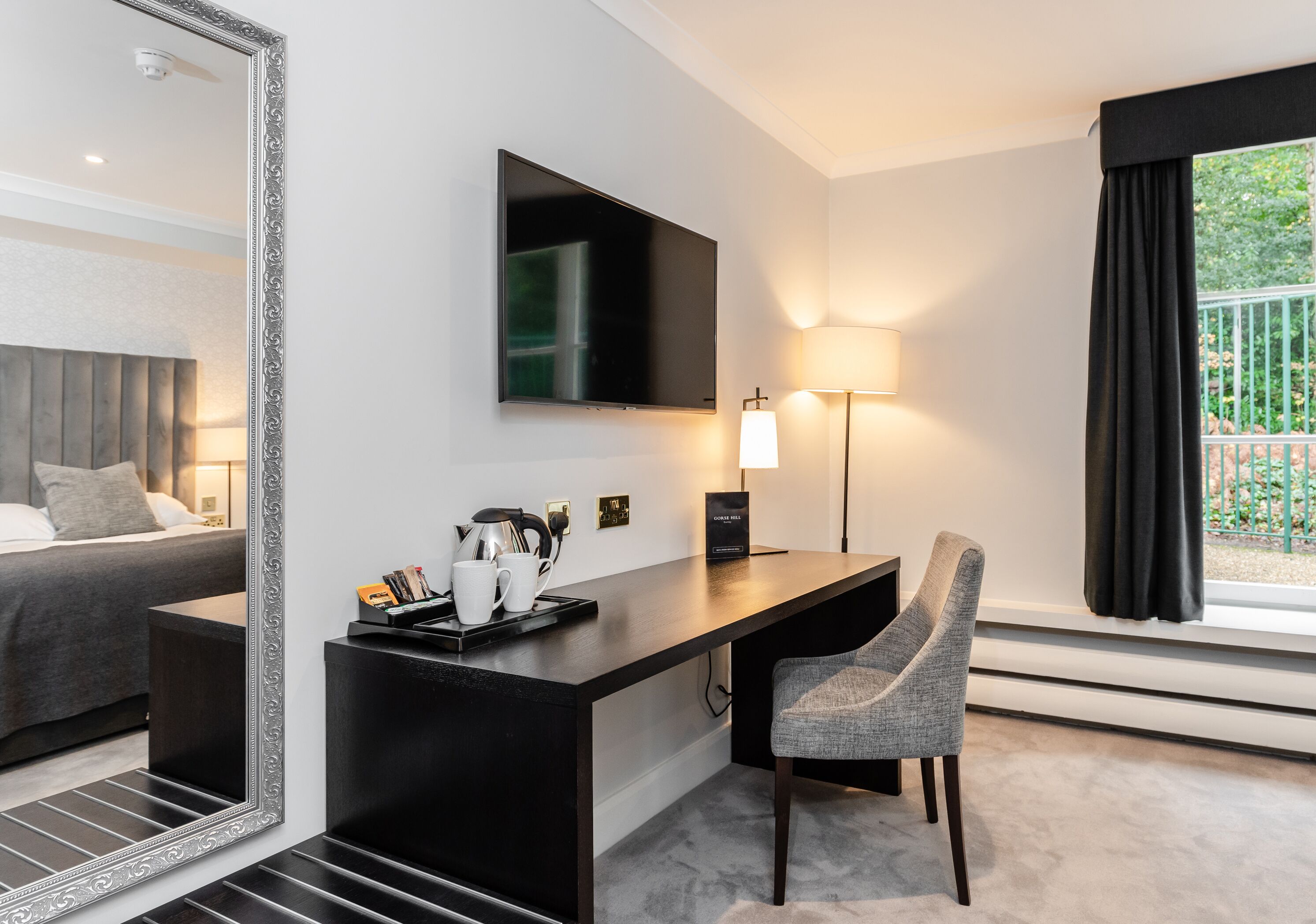A modern hotel room with a large mirror, a flat-screen TV, a desk with a chair, a bed, and a window with a view of greenery.