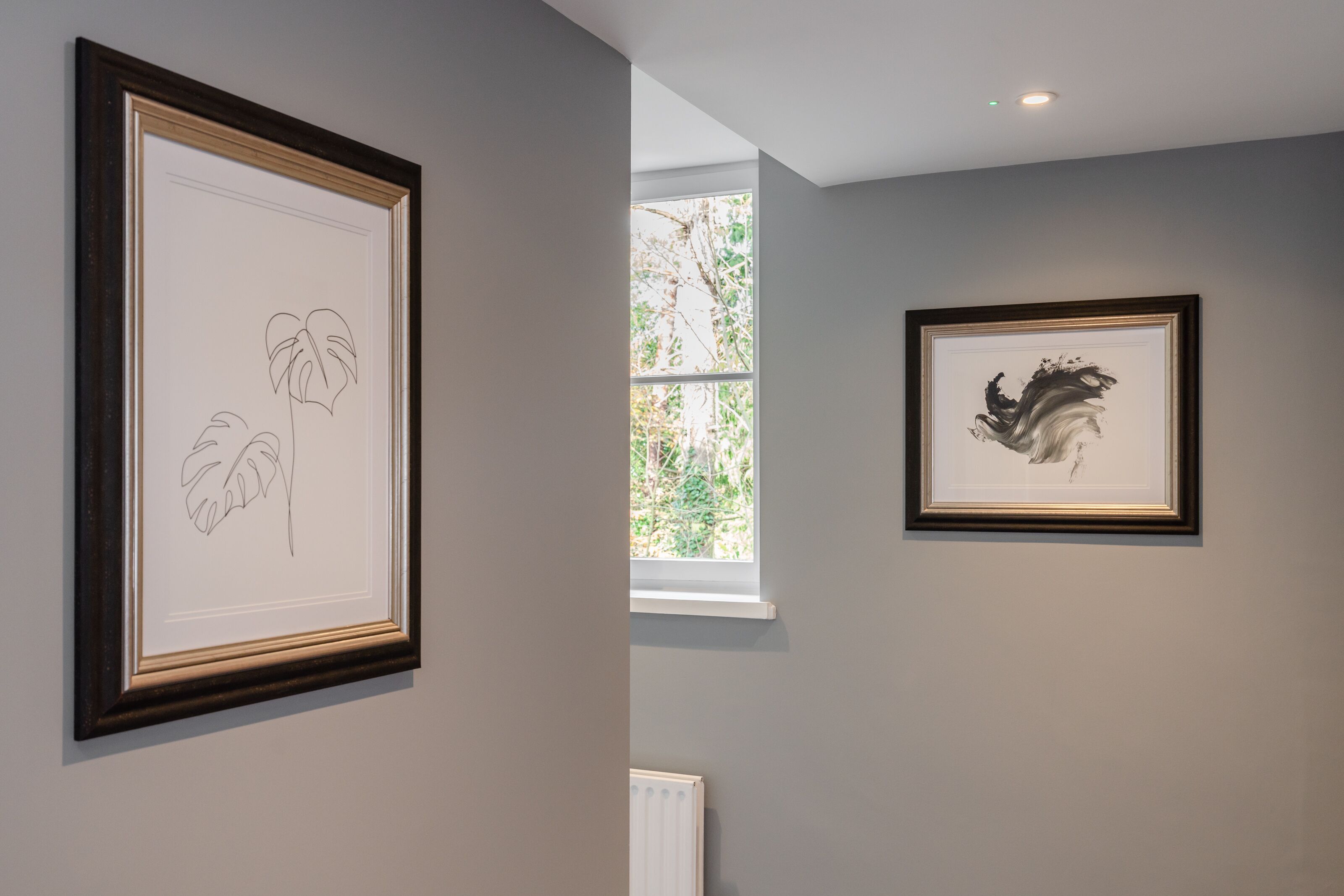 A gallery with framed artworks on the walls, a window with a view of greenery, and a radiator below the window.