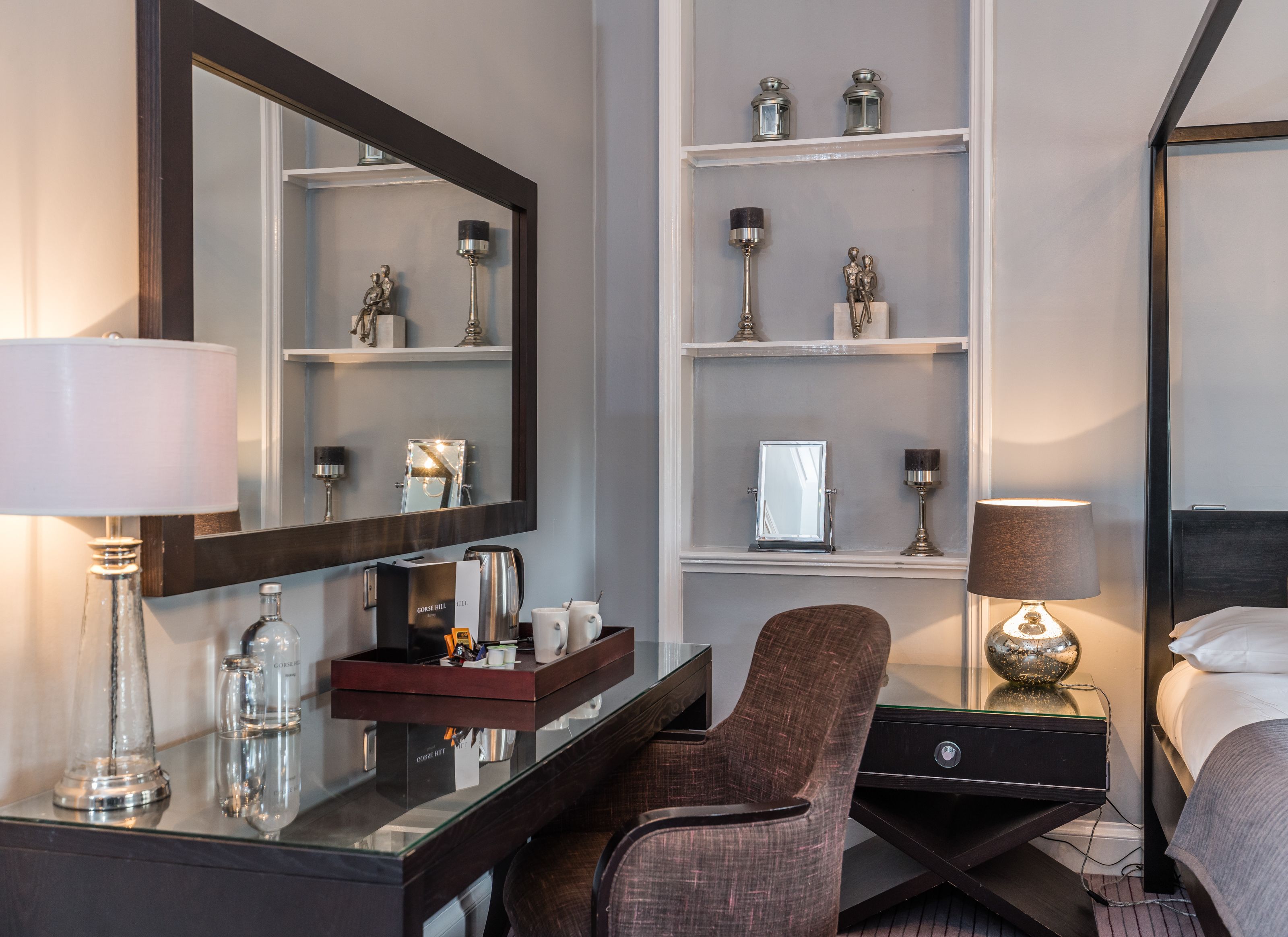 A hotel room with a large mirror, a desk with a coffee maker, a chair, a bedside table with a lamp, and shelves with decorative items.
