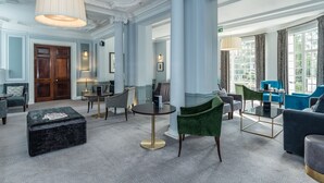A spacious lobby with a grand chandelier, elegant columns, and a mix of seating options including a plush green armchair and a blue sofa. - Gorse Hill (Woking)