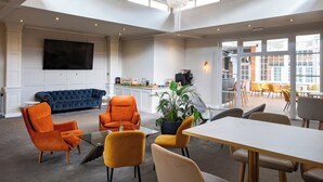 En rummelig stue med en stor lysekrone, en blå sofa, orange lænestole, et spisebord og et tekøkken med et stort fjernsyn.