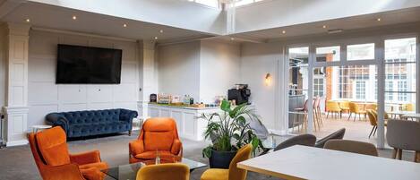 A spacious room with a large chandelier, a blue sofa, orange chairs, a dining table, and a kitchen area with a large TV.