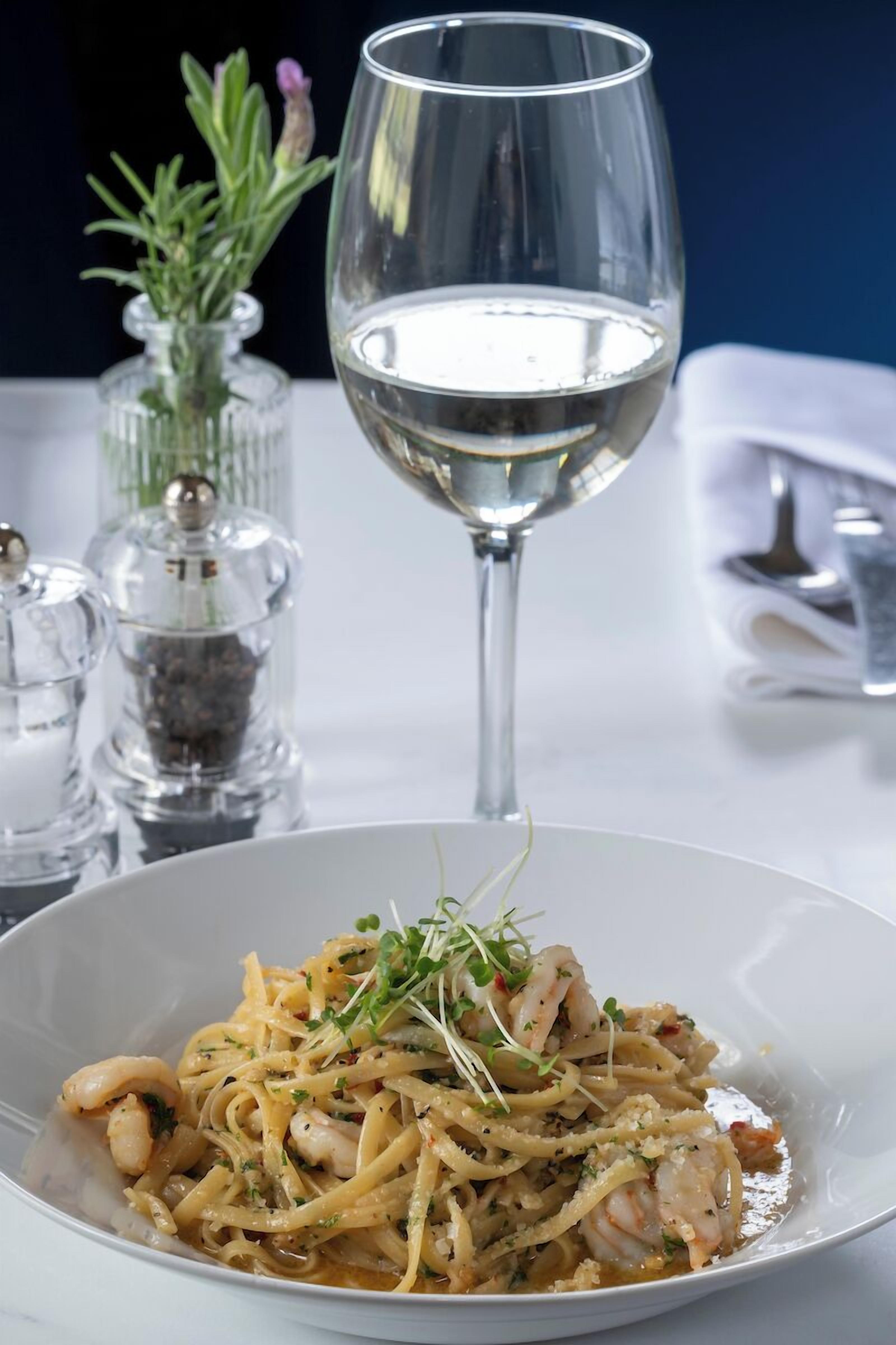 A dining table with a white plate of pasta, a glass of white wine, and a vase with green herbs.