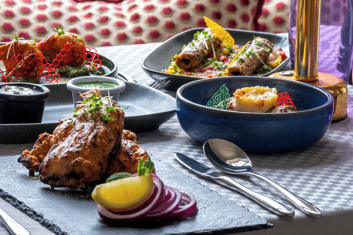 Una mesa de comedor con diversos platos, incluyendo pollo a la parrilla, tacos y un bol de comida, junto con salsas y cubiertos.